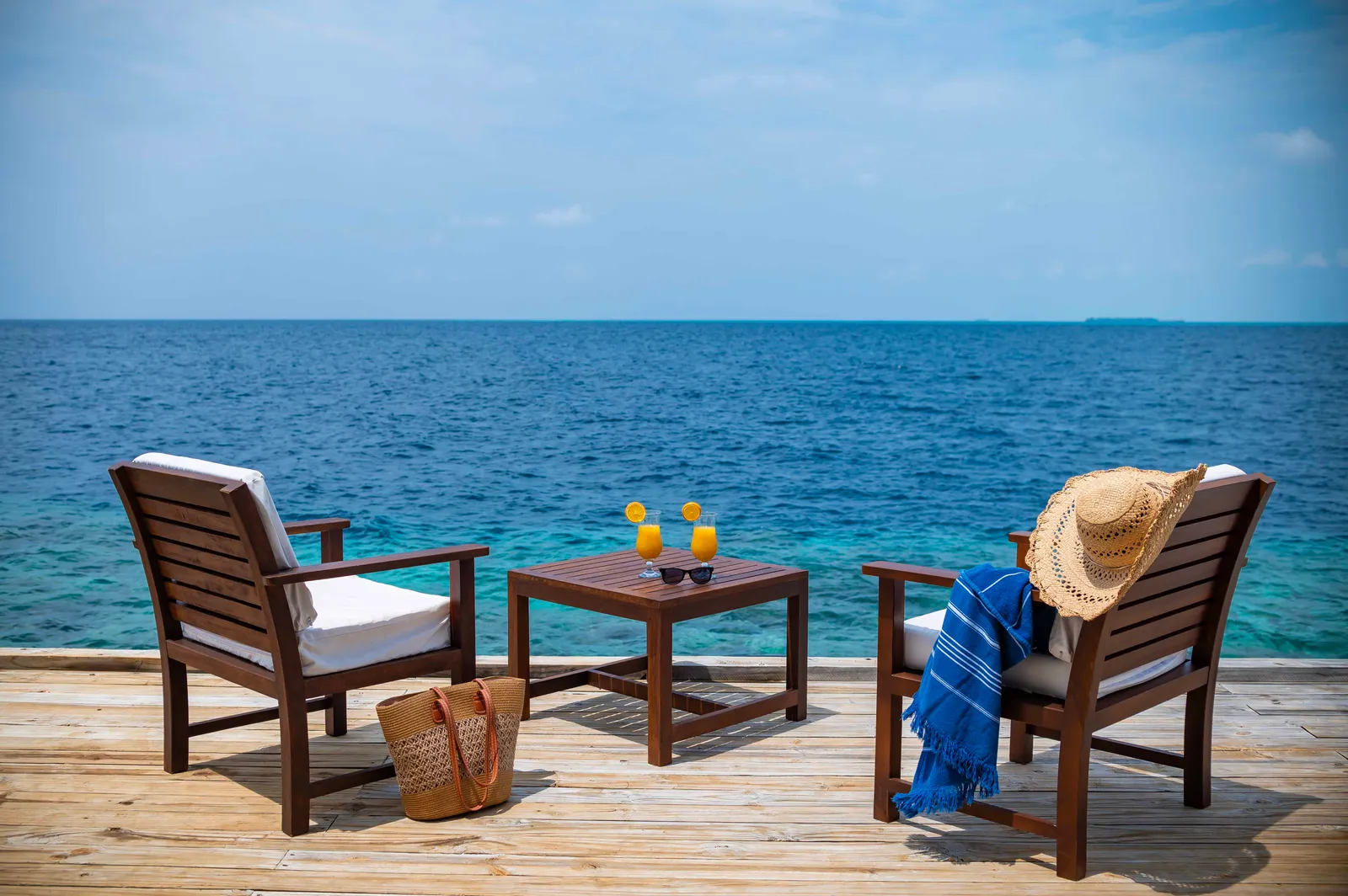A water villa’s deck: 2 wooden armchairs, a small table with 2 juices and the open sea ahead.