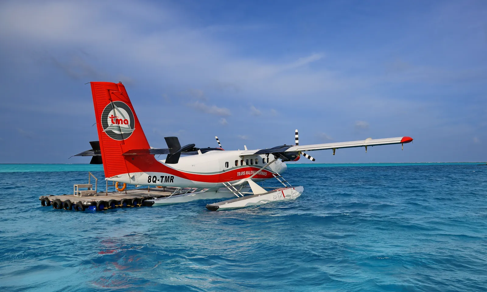 A red and white seaplane moored at the floating platform out at sea, off the island.