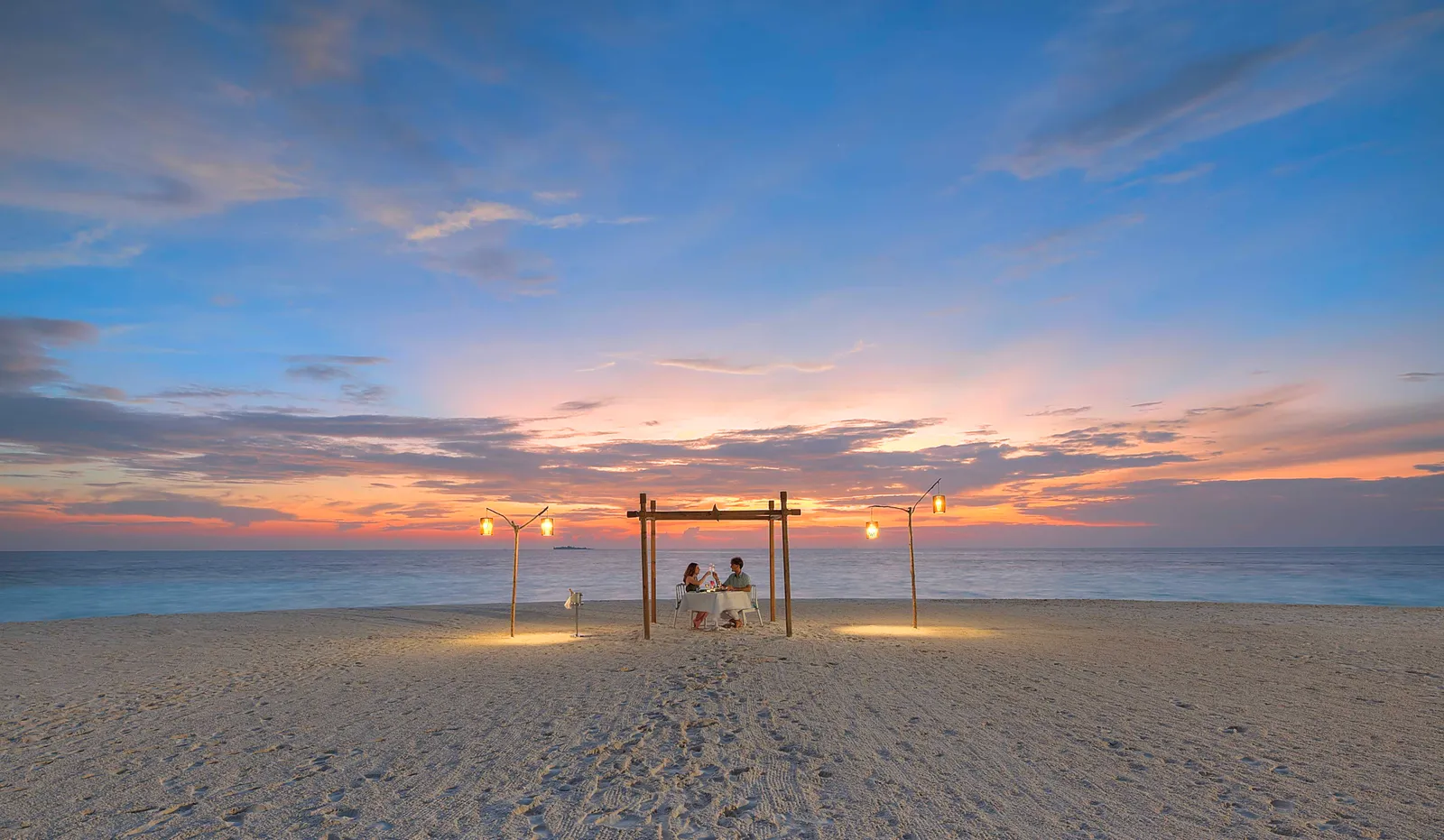 A swing on the beach at sunset, 2 people seated between torches lit on the sand.