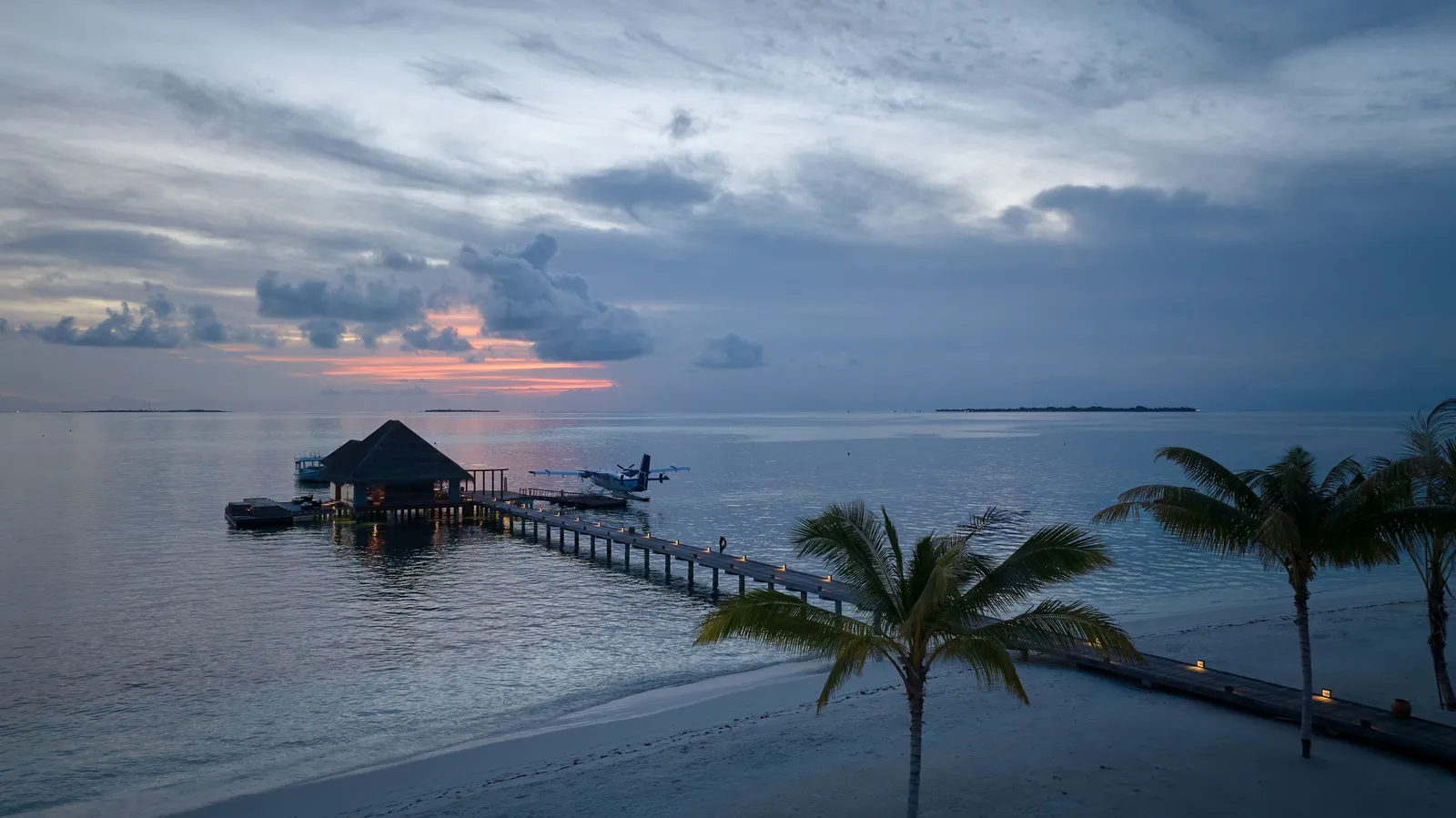 The arrival jetty at dusk: the thatched pavilion over the water, a seaplane moored alongside and palms on the beach