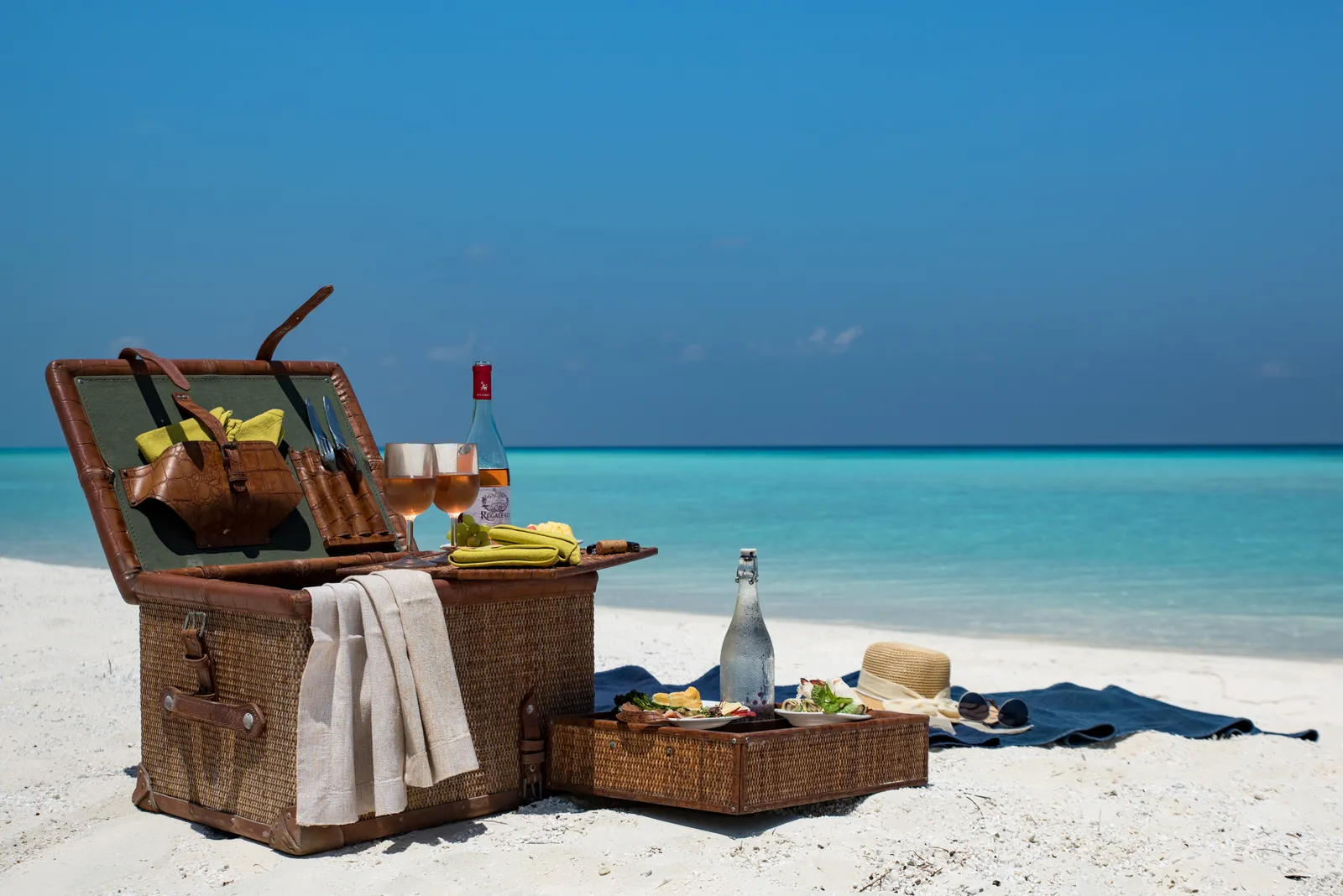 A wicker picnic hamper, wine and glasses on the sand of Masleggihuraa, facing the sea