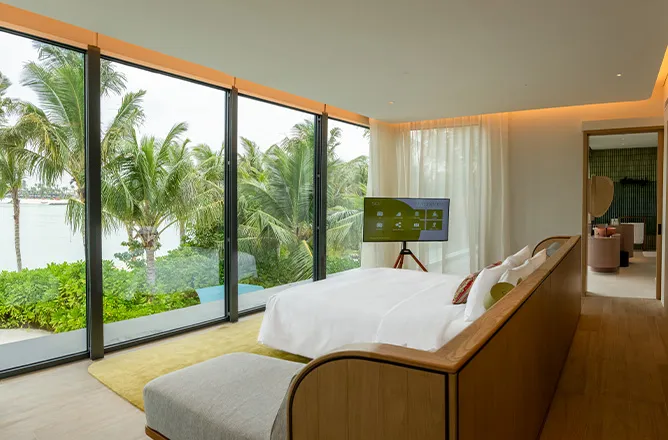 Upstairs bedroom behind large glass windows with palm and lagoon outlook; bed,TV,wood floor.