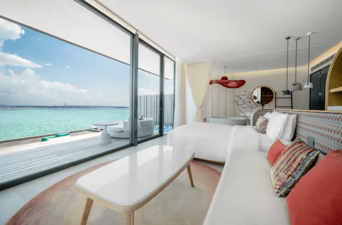 Bright white bedroom/lounge with ocean through broad glass doors; deck and loungers.