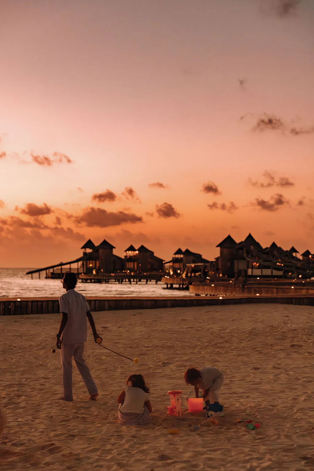 3 children on the beach at sunset, the lit water villas behind them