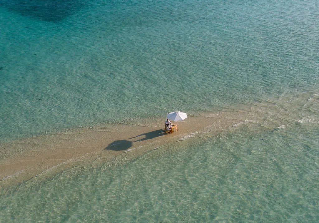 A table under a white parasol on a strip of sand in the open sea