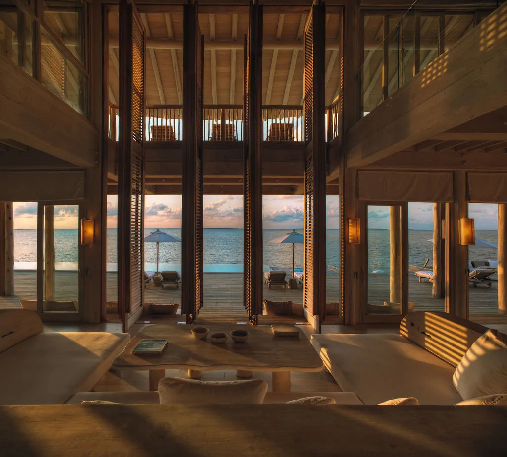 The double-height living room of a water villa, glass doors open to the deck at sunset