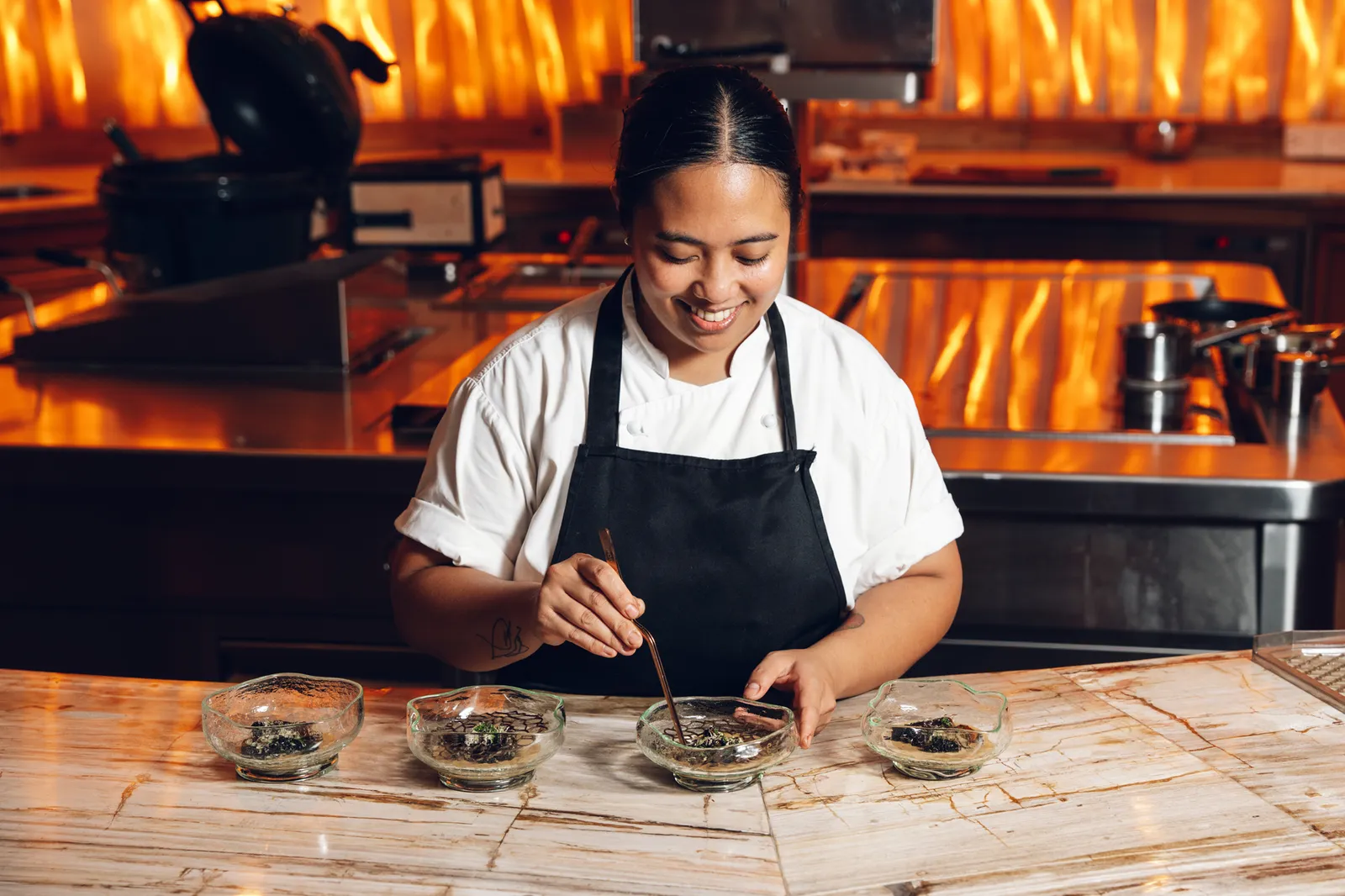 A chef plating 4 small bowls on a marble counter, in front of the kitchen’s flames.