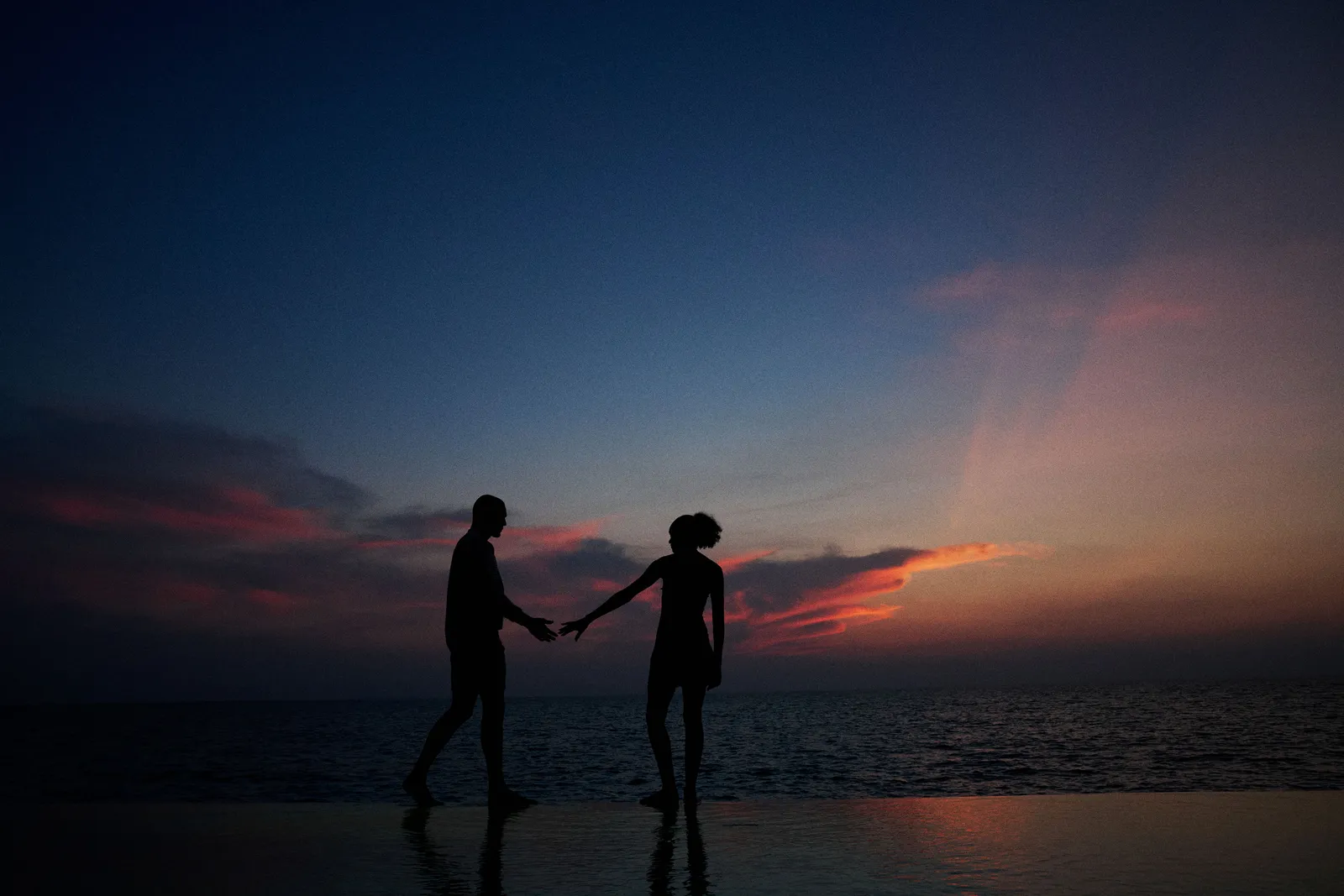2 people holding hands, silhouetted at the water’s edge after sunset.