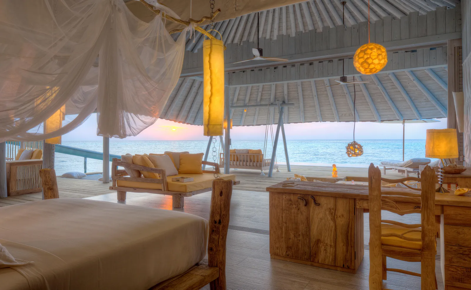 The bedroom of an overwater villa, with the bed under its net, a desk and the sea beyond the glass.