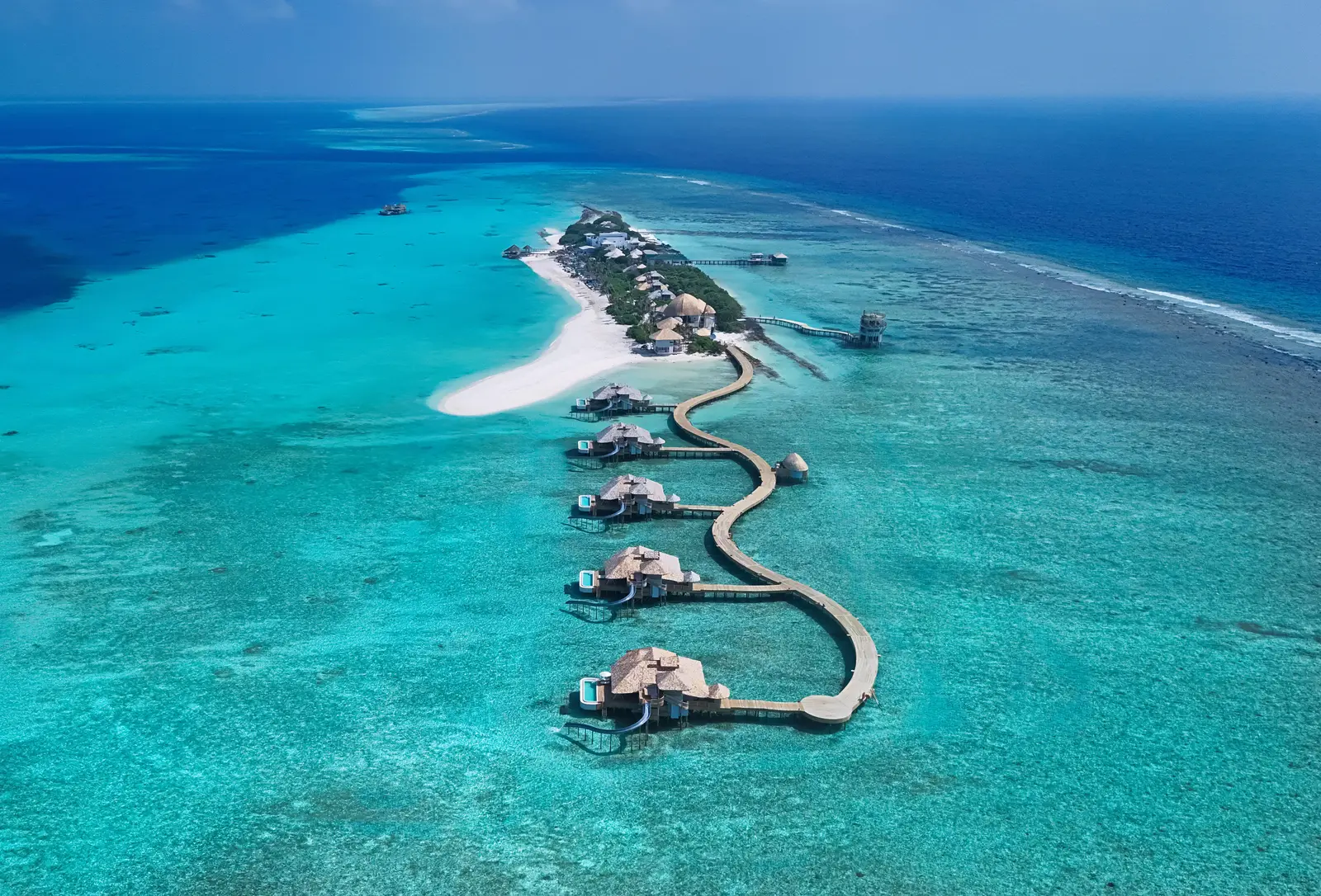 The island from above: the spit of white sand, the winding jetty and the overwater villas in a row on the lagoon.