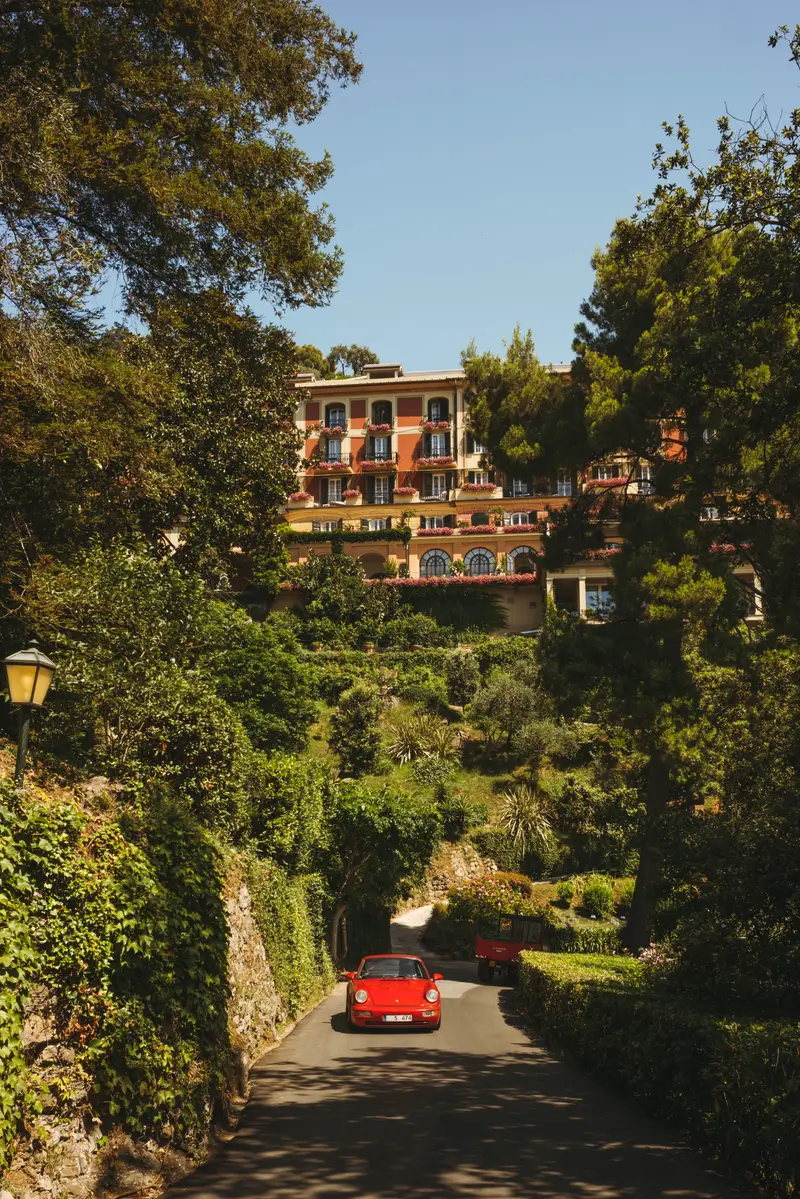 The hotel’s pink façade at the top of the drive, among the pines, a red vintage car on the road.