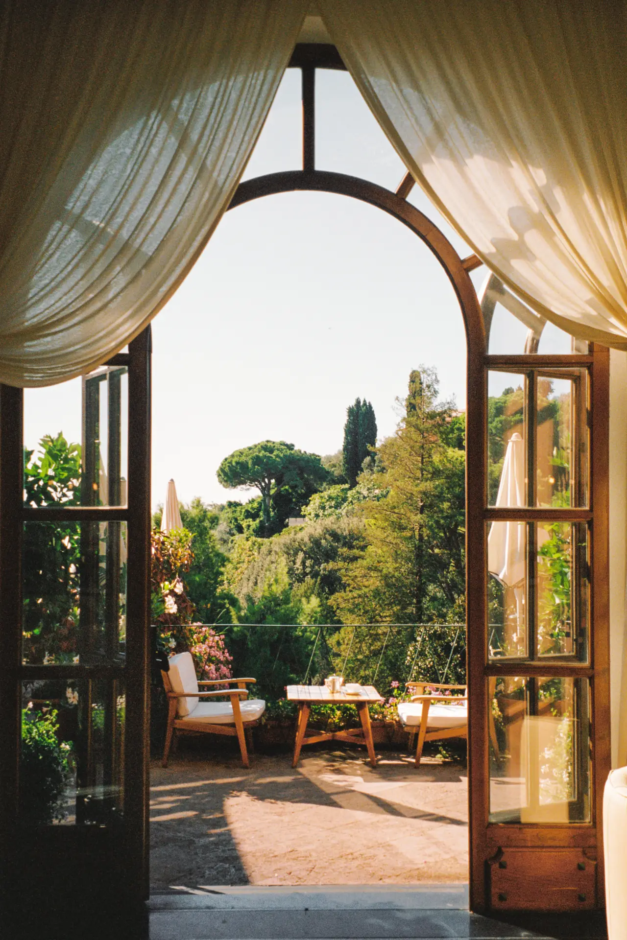 A garden terrace seen through a curtained arched door, with armchairs and umbrella pines.