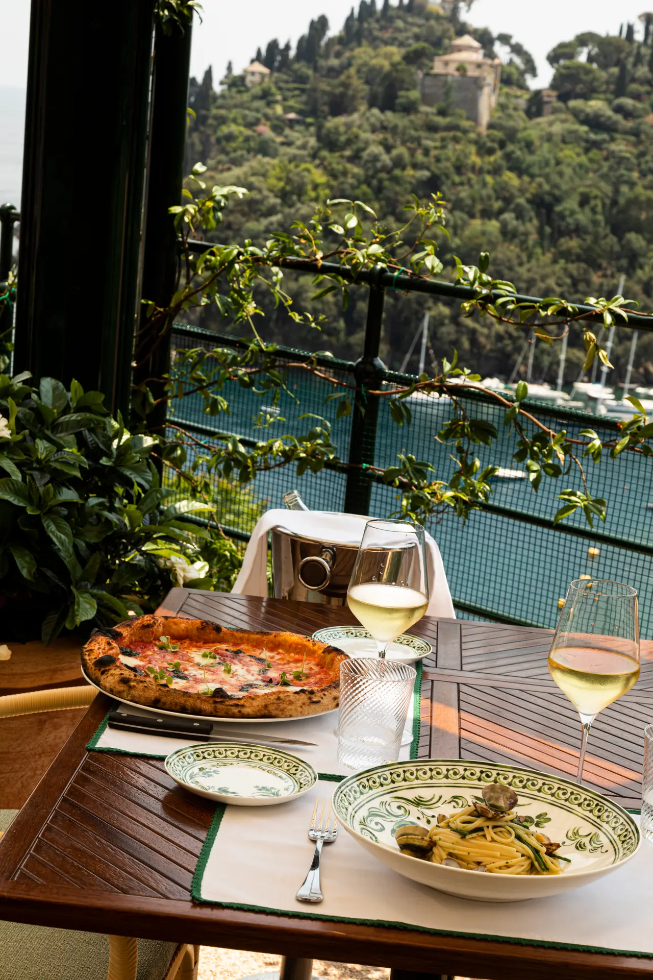 A pizza, a plate of pasta and 2 glasses of white wine on a Splendido Grill table, the harbour below.