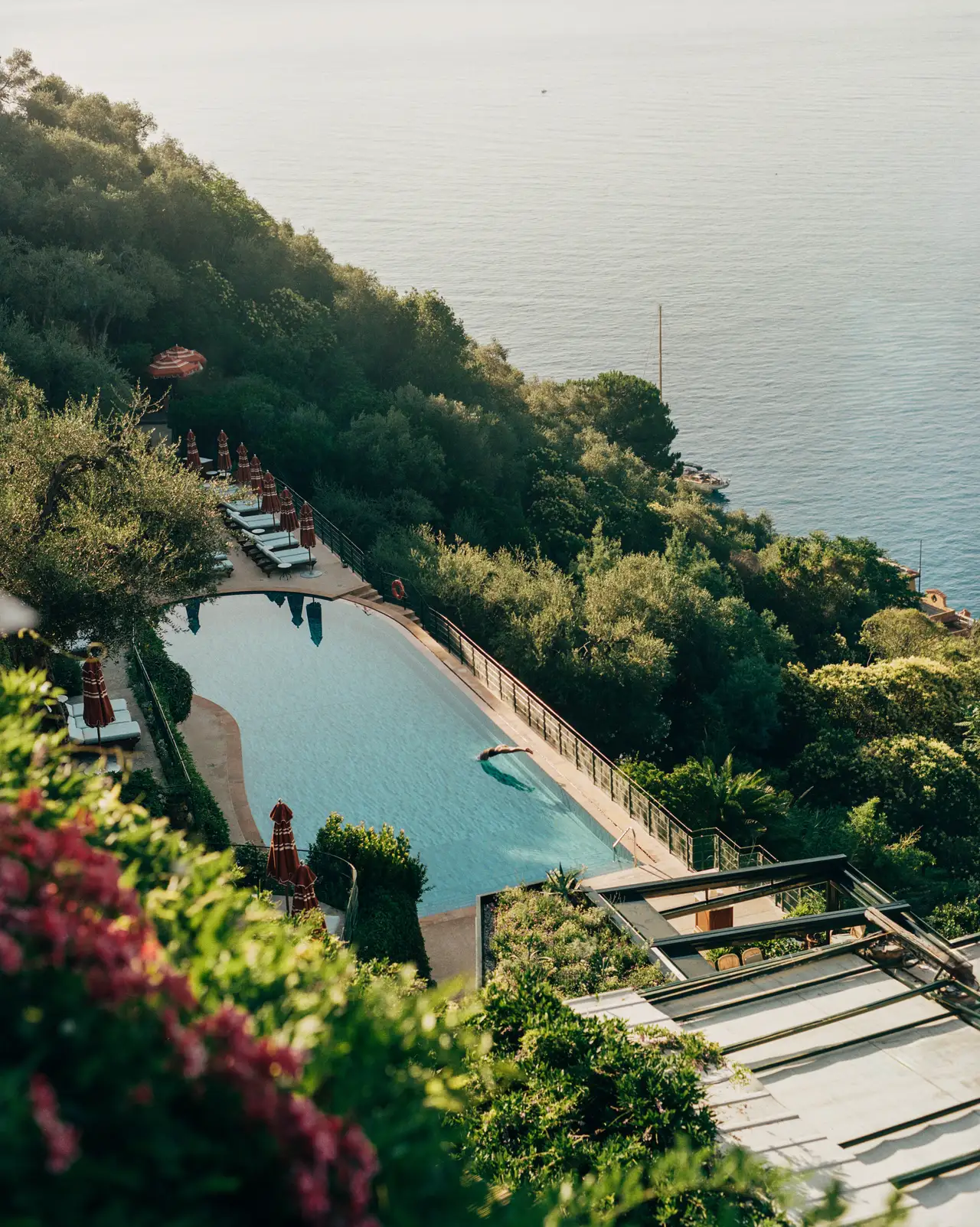 Splendido’s pool from above, long between the terraced gardens, with red parasols and the sea below.