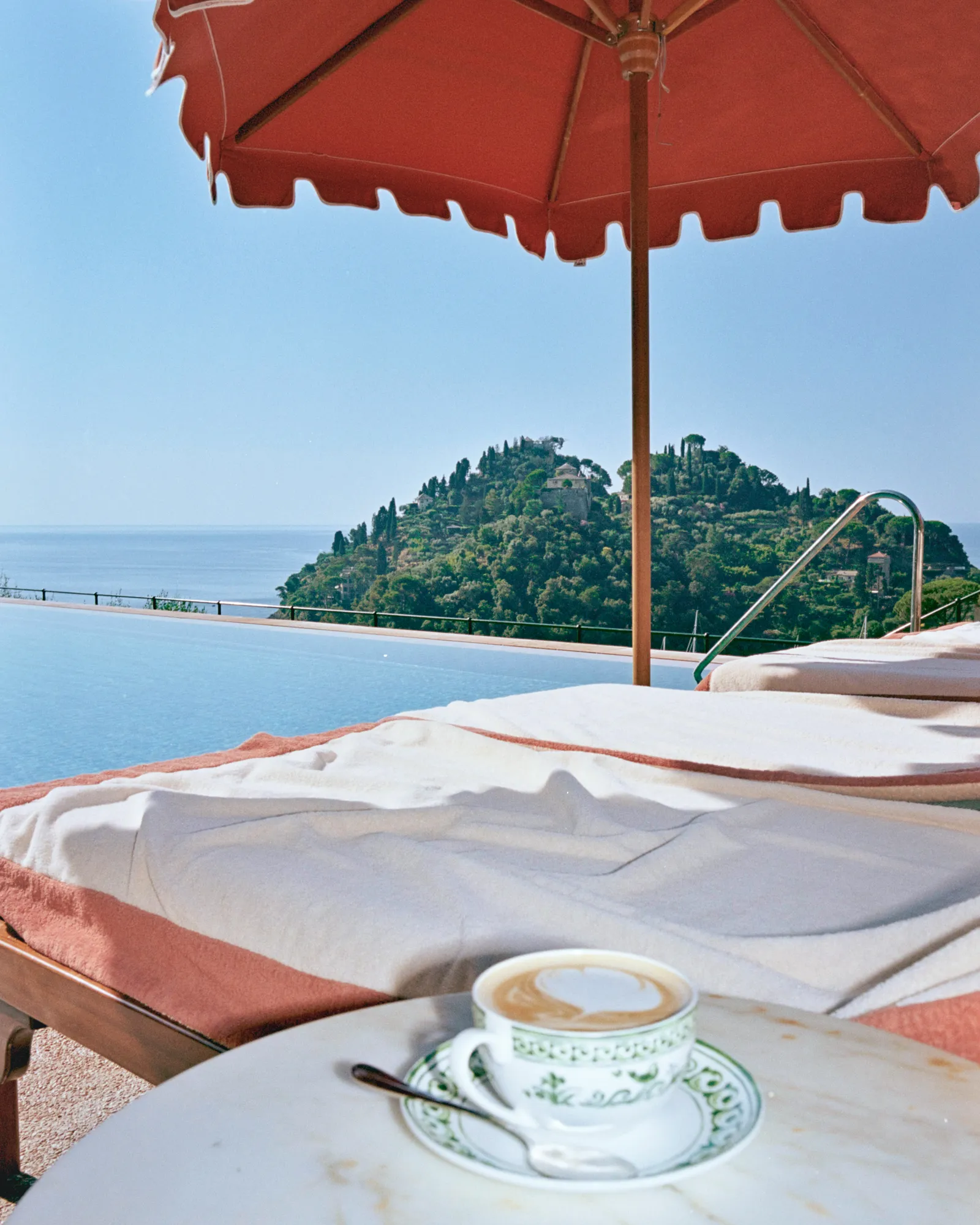 The pool’s infinity edge under a red parasol, a cappuccino on the side table and Portofino’s headland opposite.