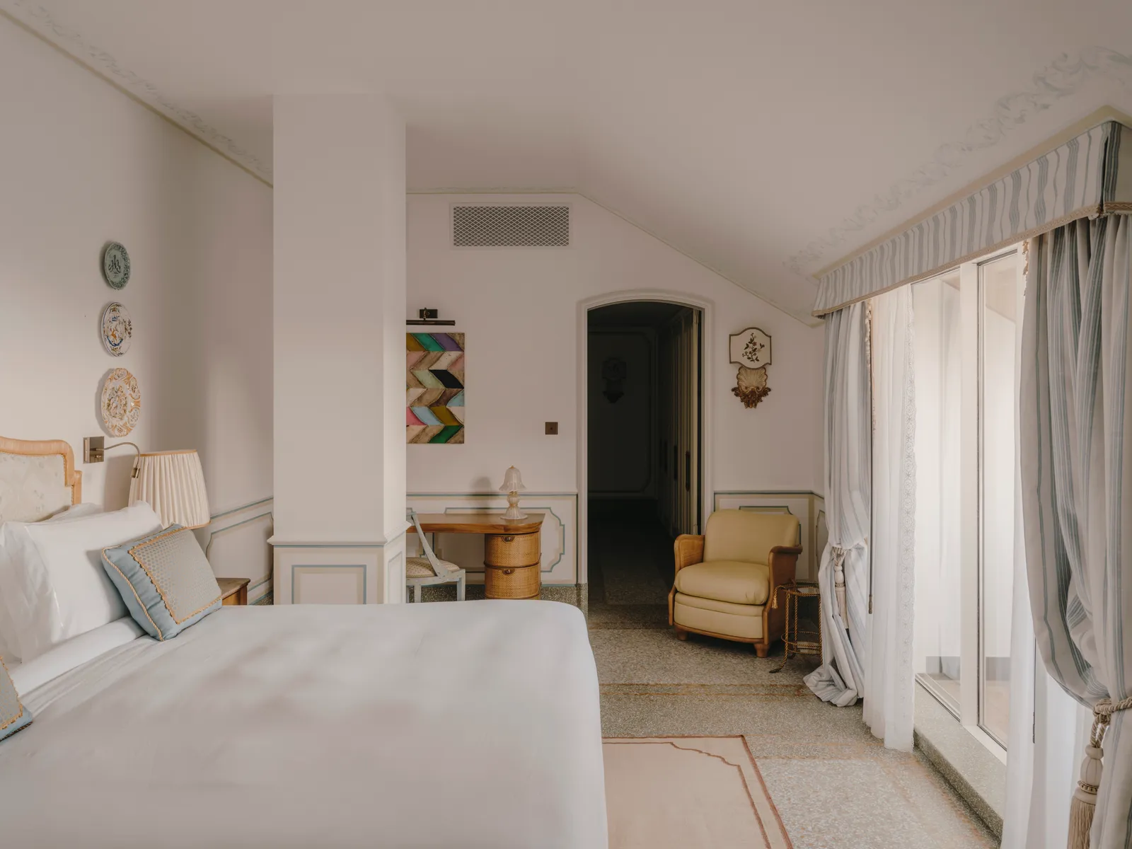 A Portofino Suite bedroom: the bed, 3 decorative plates on the wall and curtains at the French window.