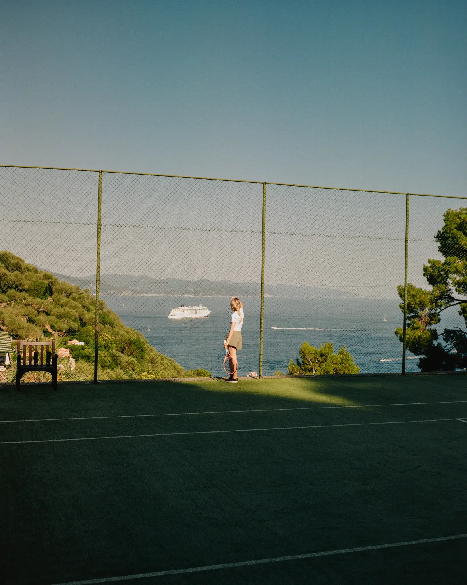 A player on the hillside tennis court, a white ship on the sea beyond the fence.