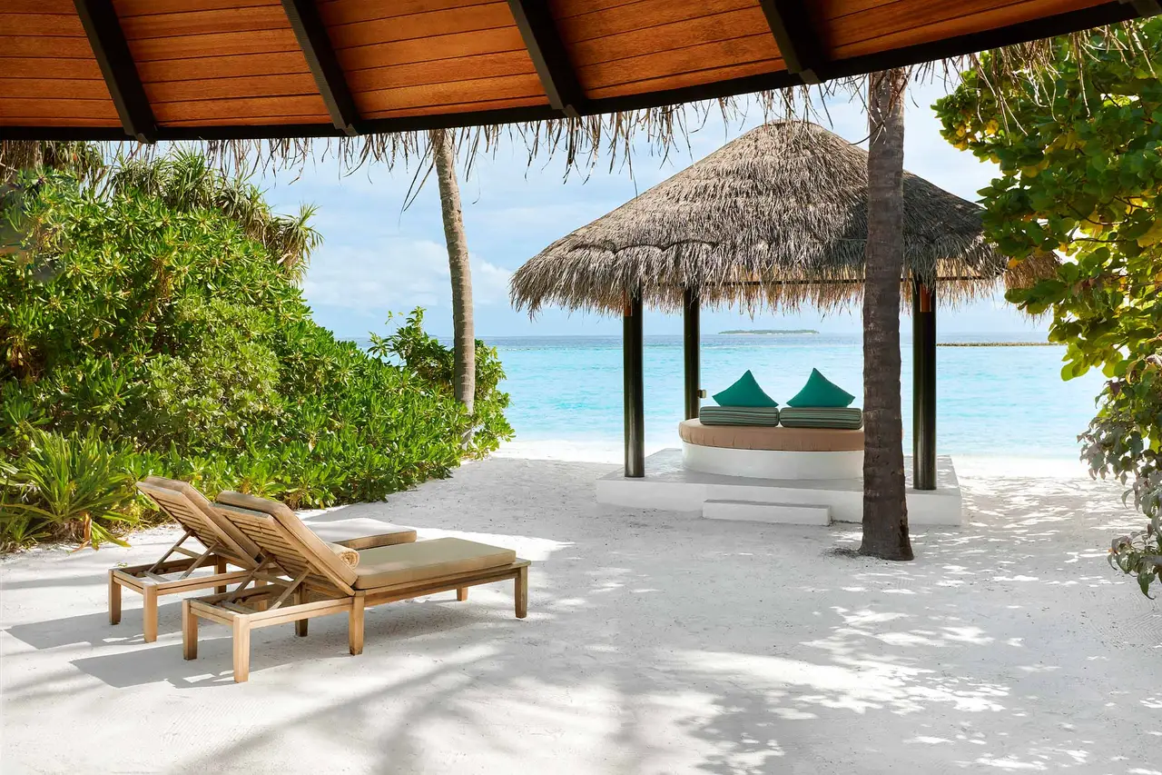 Under a beach villa’s porch: loungers, the thatched sala and the sea between the trees.