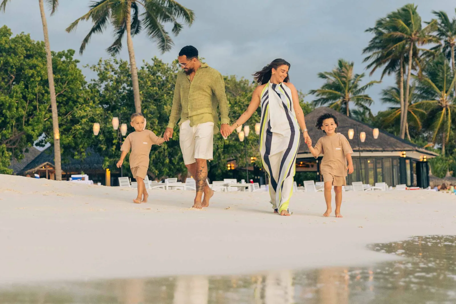 A family walks along the beach at sunset, the children ahead, lanterns lit among the trees.