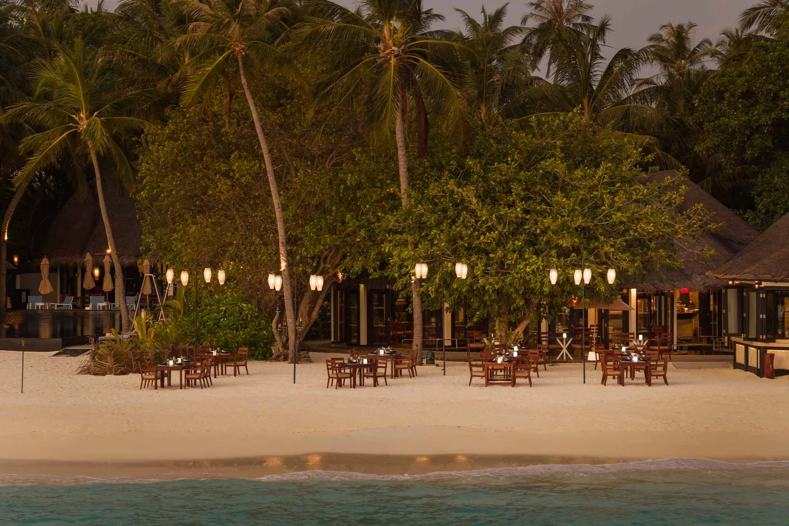 Islander’s Grill at dusk: tables on the sand, lanterns lit among the palms and the sea a few steps away.