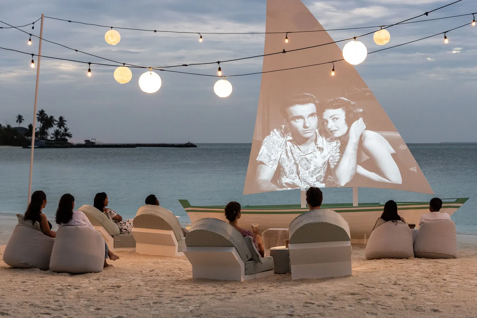 The beach cinema: guests on beanbags facing the sail used as a screen, lanterns strung above.