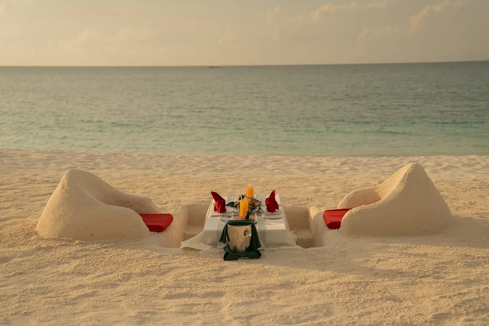 A table for 2 dug into the sand, with sculpted seats and flowers, facing the sea at sunset.