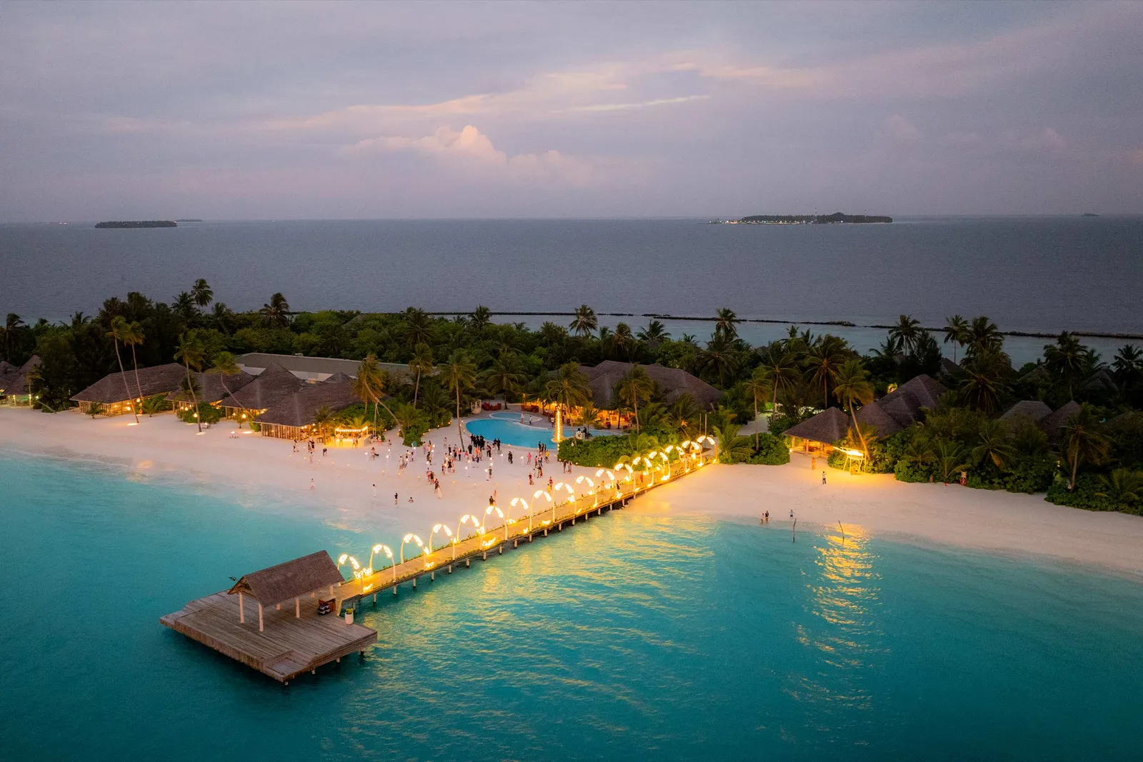 The arrival jetty lit up at dusk, the beach with guests on it and the island’s palms under a violet sky.