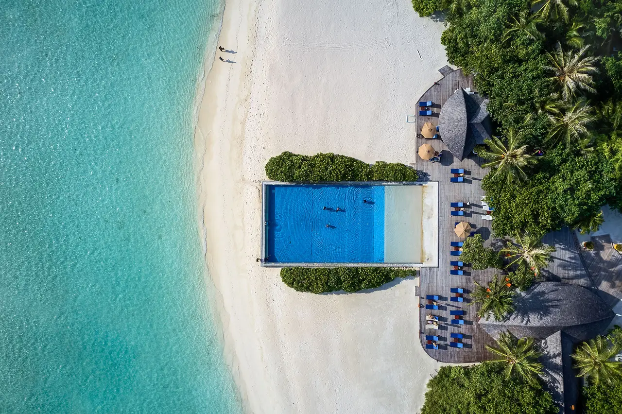 A blue pool between the beach and the palms, seen from above