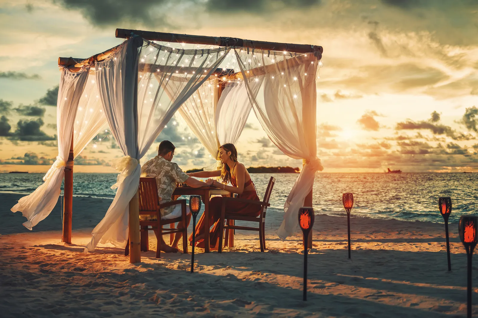 Dinner for 2 under a draped pavilion on the beach at sunset, with torches lit in the sand.