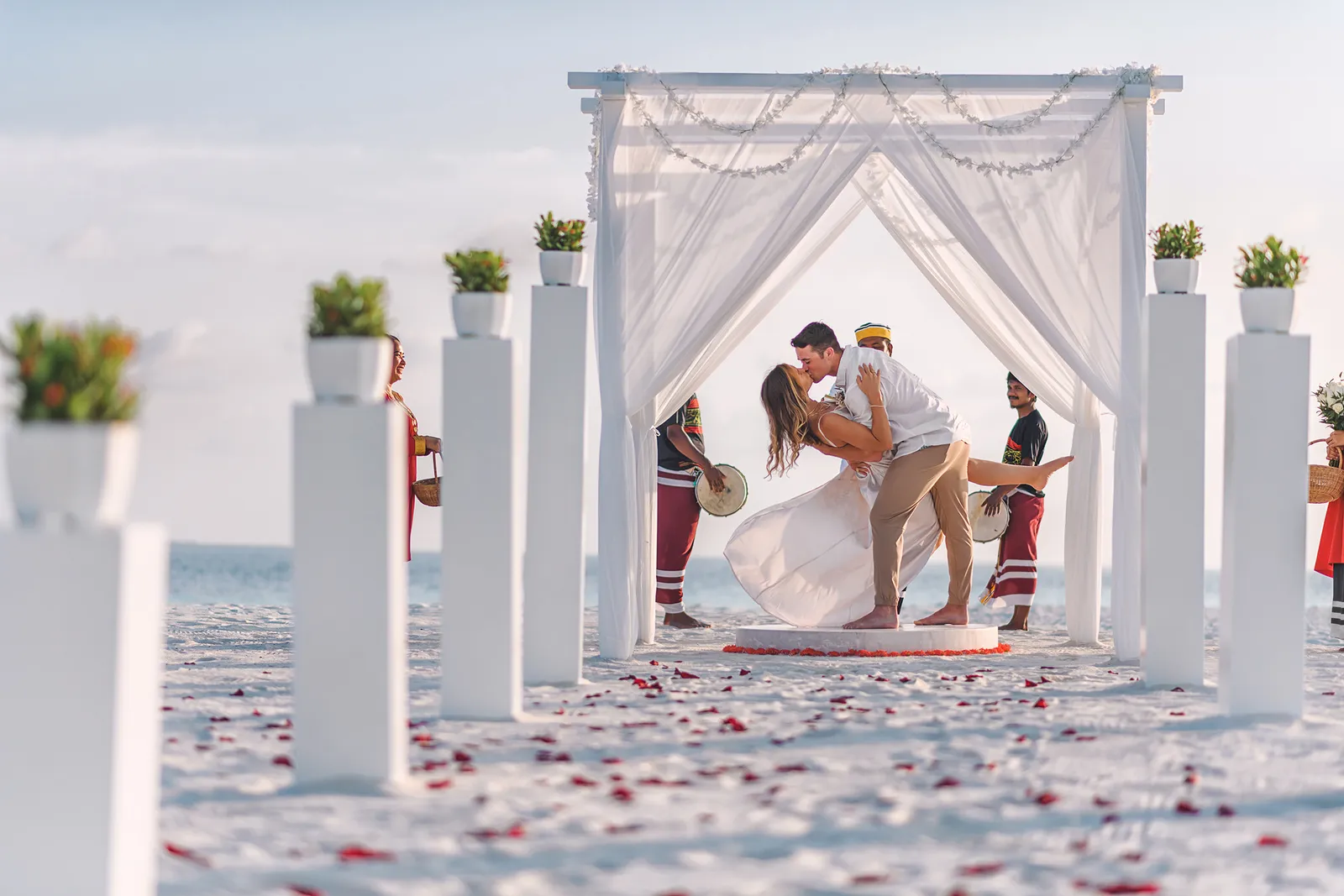 A couple kissing under the white ceremony arch on the beach, between columns topped with plants and petals.