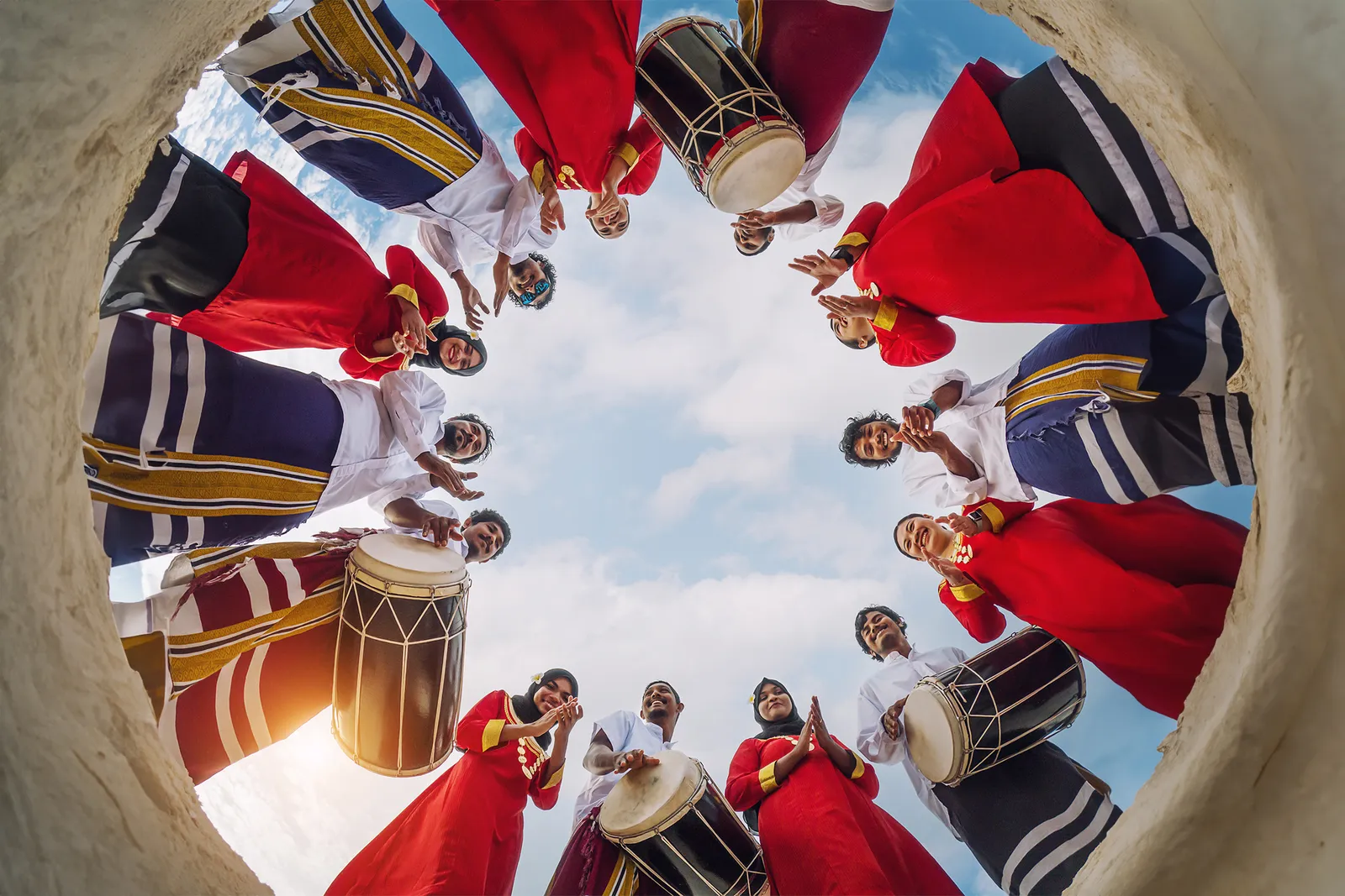 The bodu beru players seen from below, in a circle with their drums and red and white costumes, against the sky.
