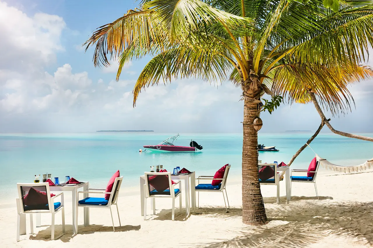 Well Done’s tables on the sand beneath a palm, with the shallow, clear water and a boat on the lagoon.