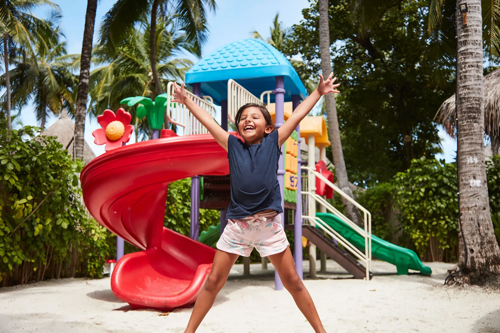 A girl jumping for joy in front of the red slide in the Turtle Kidz Club playground, among the palms.