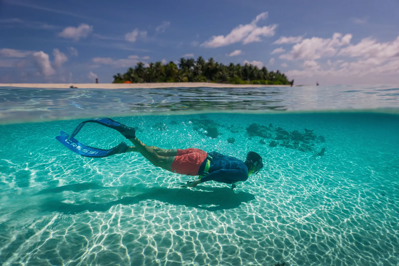 A young man in fins swimming underwater above the pale seabed, with the green island on the horizon.