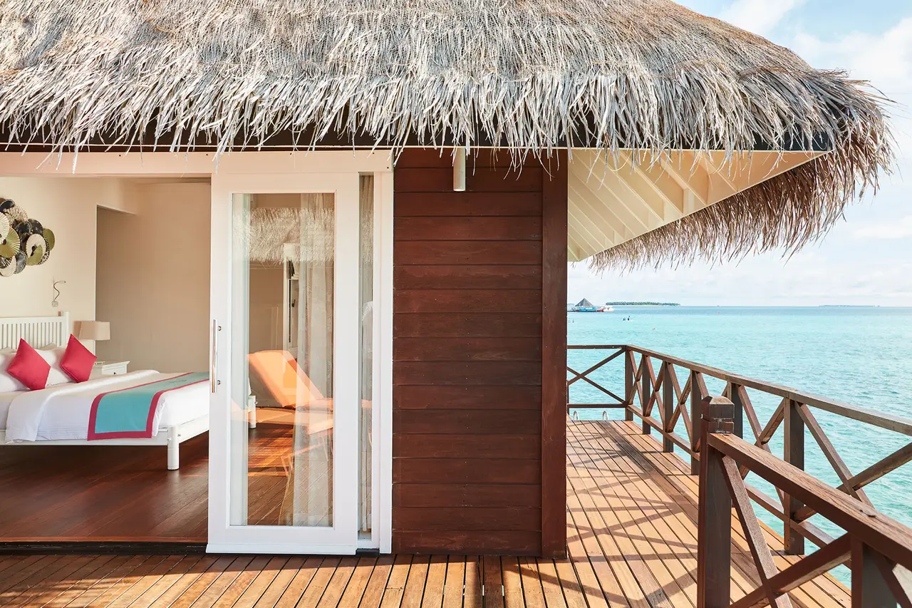 A Sunset Reef Villa’s bedroom opening onto the deck above the lagoon, under the thatched roof.