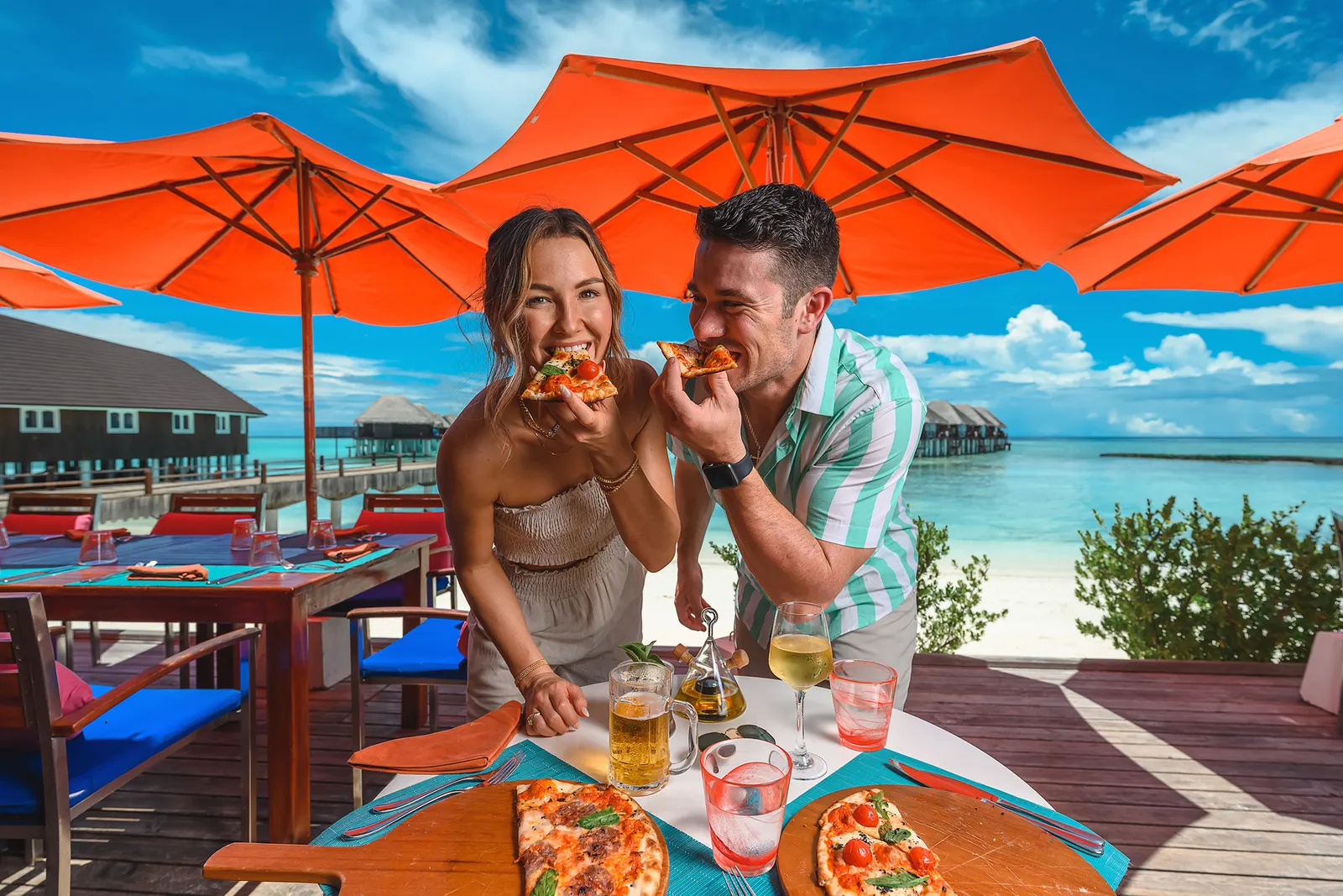 A couple biting into pizza on The Spice’s terrace, under orange parasols, with the water villas behind.