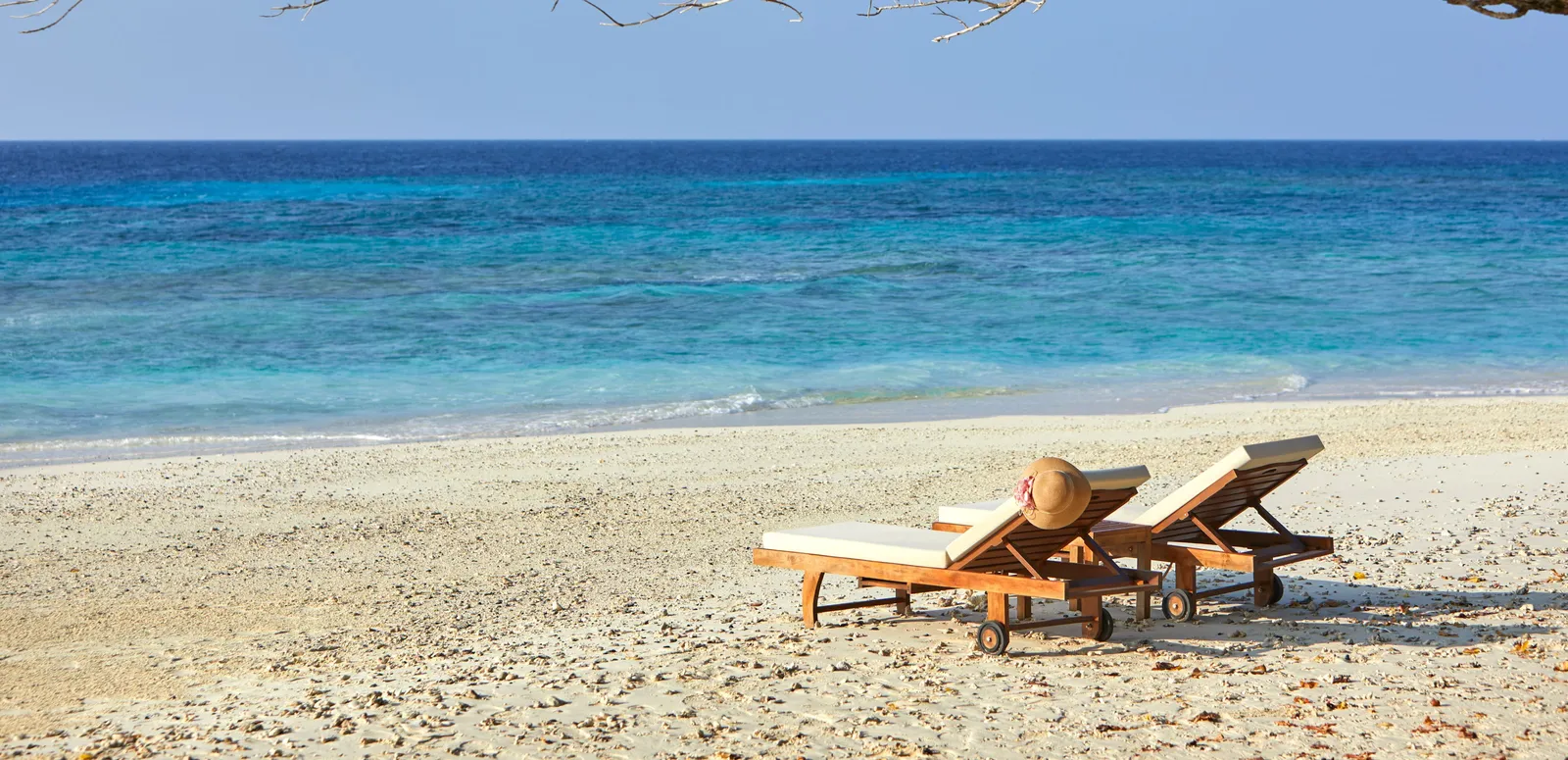 2 wooden loungers on the white beach under a branch, the turquoise sea ahead.