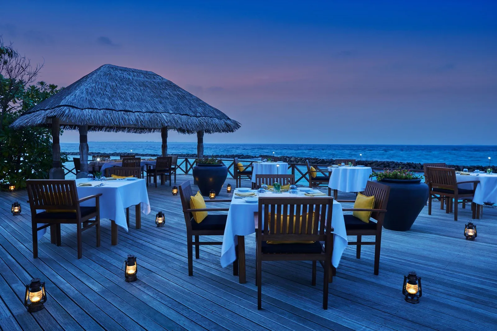 Tables laid on the wooden deck over the sea at dusk, lanterns lit beside a thatched pavilion.