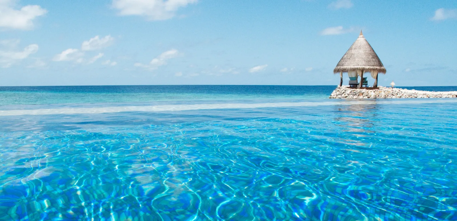 The infinity pool merging into the sea, with the thatched gazebo on the tip of the breakwater.