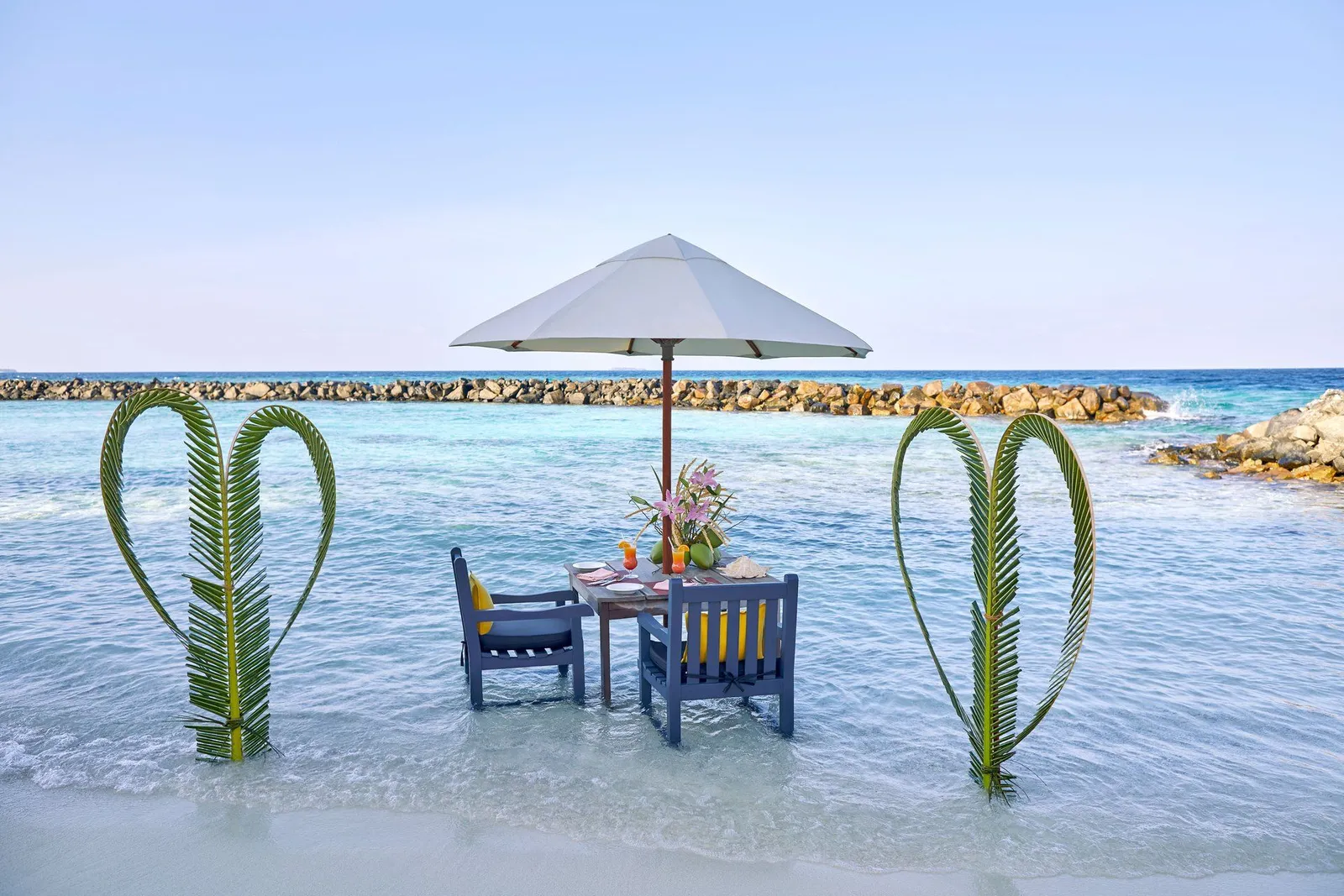 A table for 2 laid in the shallow lagoon under a parasol, between 2 woven palm fronds.