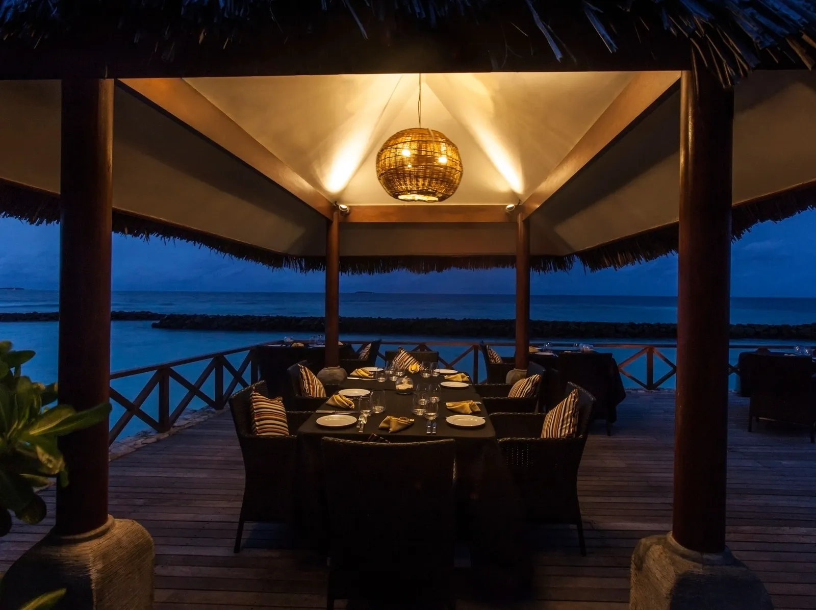 A wooden pavilion over the water laid for dinner, its lantern lit against the blue of the evening sea.