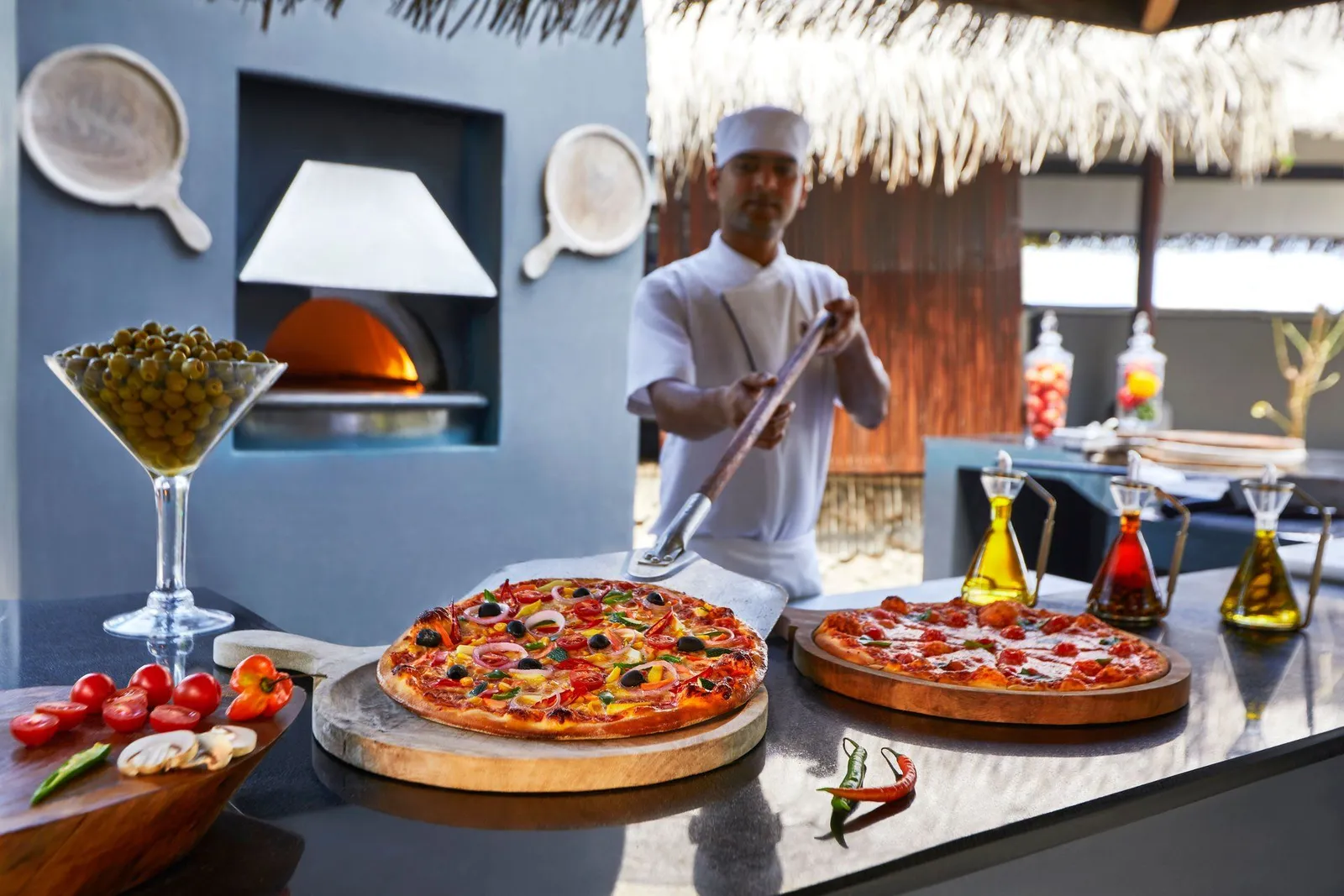 A chef drawing a pizza from the wood oven, with finished pizzas on the counter.