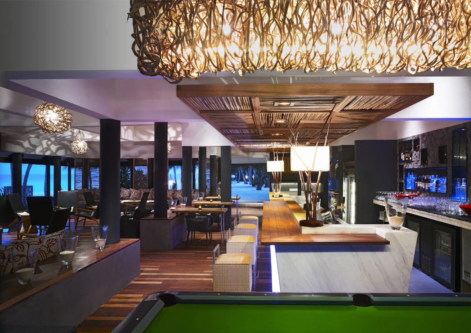 The Reef Bar at night, with its lit counter, the pool table and the chandelier of woven branches.
