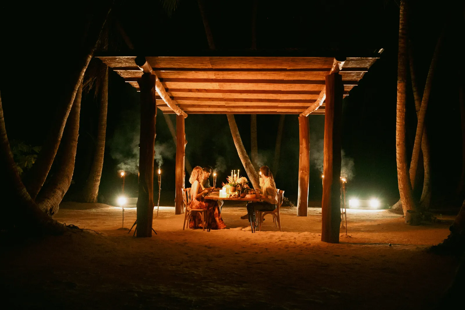 A table for 2 under a timber pavilion on the sand at night, lit by lanterns