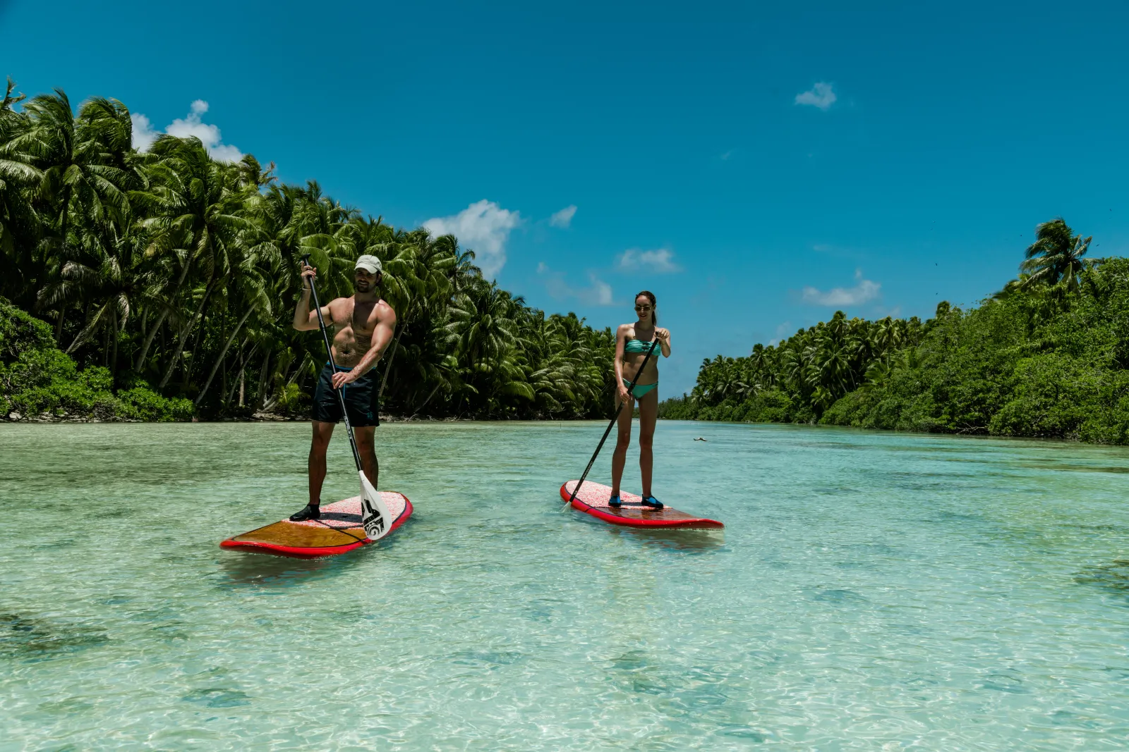 2 paddle boarders on the clear lagoon between 2 wooded motus
