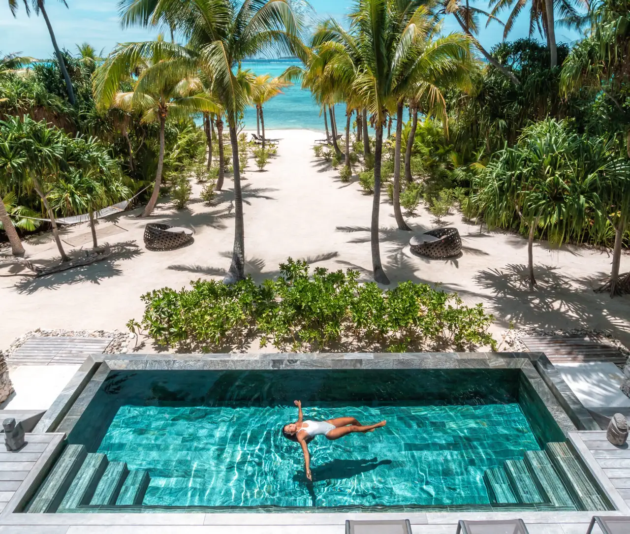 A villa’s pool from above, someone swimming, the deck and the beach beyond the palms