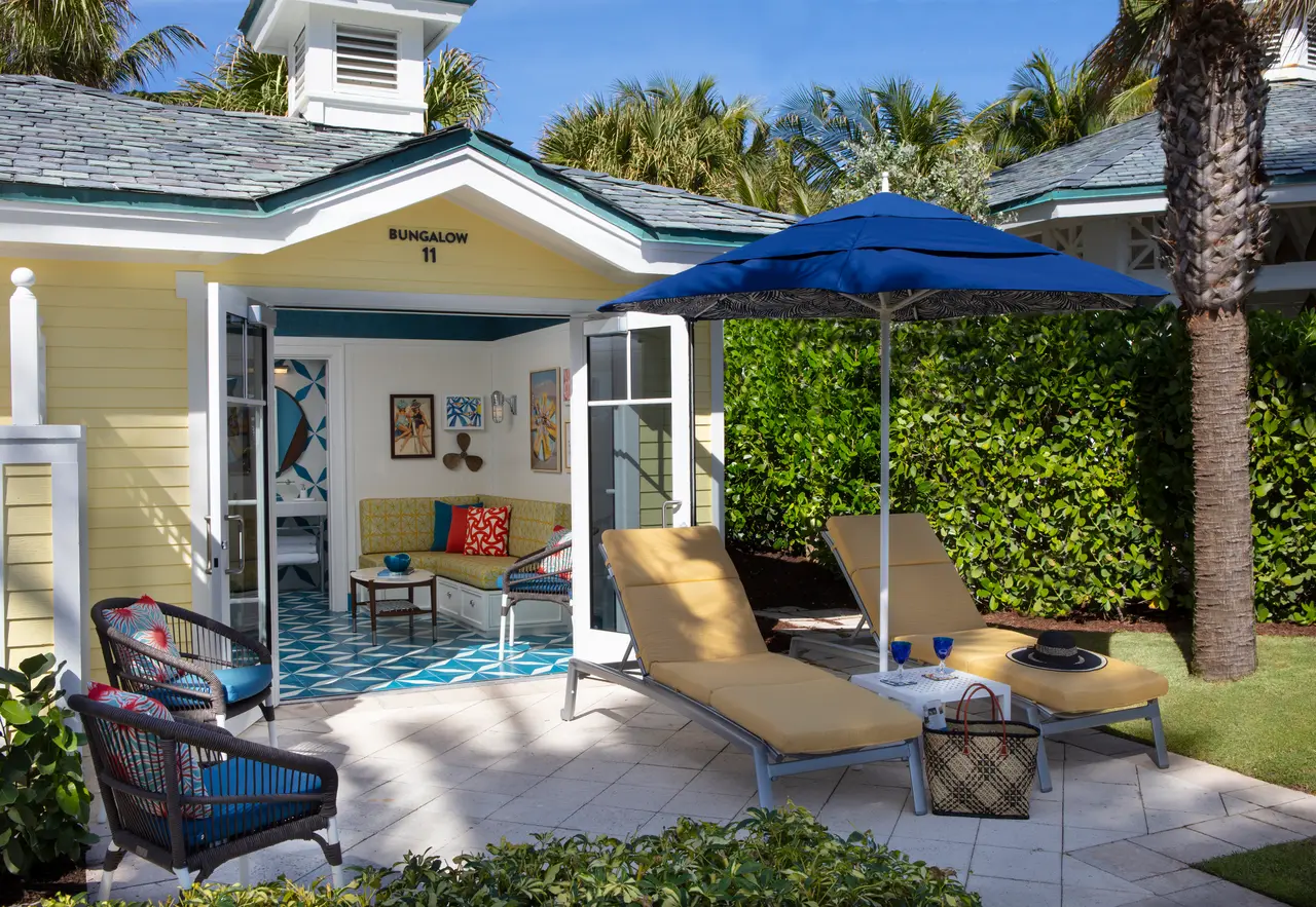 One of the poolside bungalows: the yellow cottage with its doors open onto the sitting room, 2 loungers and a blue parasol on the patio.