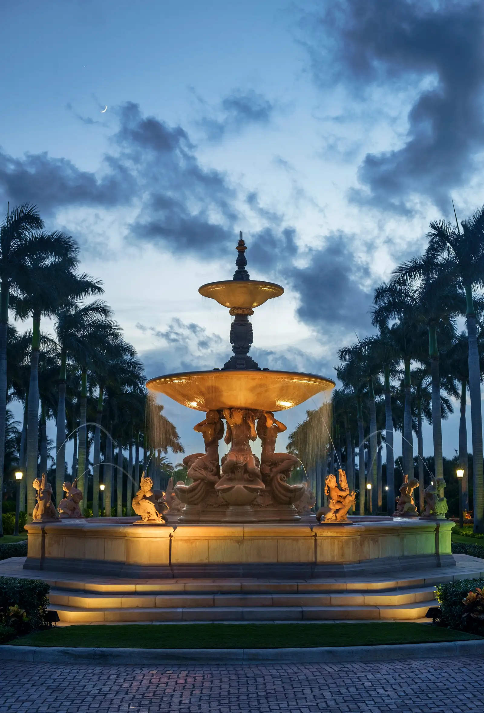 The Florentine Fountain on the main drive, lit at dusk between 2 rows of royal palms.