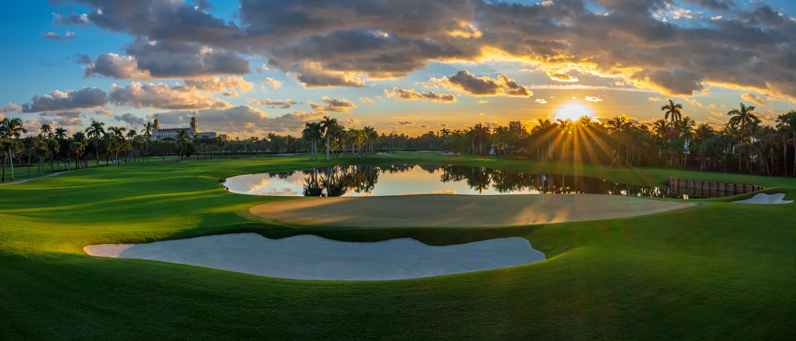 The golf course at sunset: a pale sand bunker, a pond mirroring the clouds and the sun low between the palms.