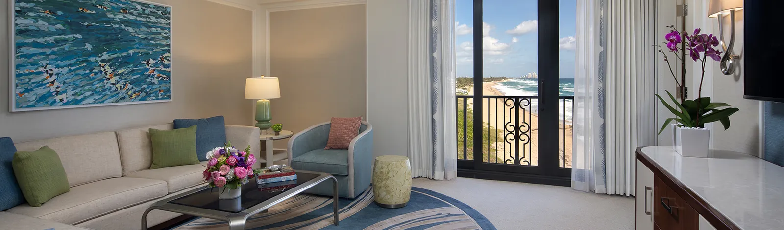 An Atlantic Suite’s sitting room: the sofa with green and blue cushions, a seascape on the wall and the Juliet balcony over the beach.